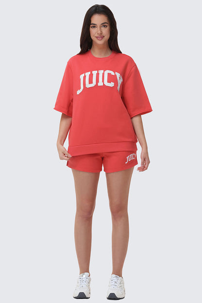 A woman faces forward, smiling, in a relaxed fit red Collegiate Sweatshirt with matching shorts and white sneakers against a plain light background.