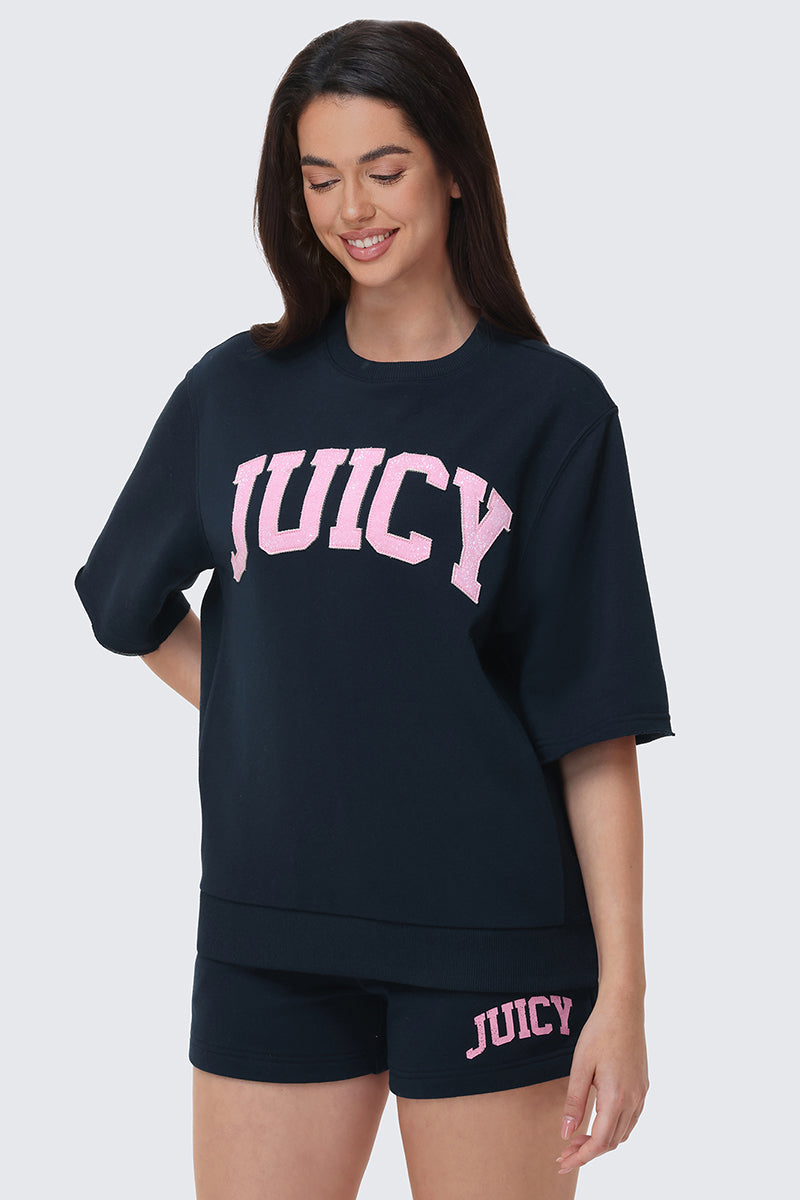 A woman smiles with her eyes closed, wearing a collegiate-style navy blue Collegiate Sweatshirt with JUICY in large pink letters and matching navy blue shorts featuring JUICY in smaller pink letters on the left leg.