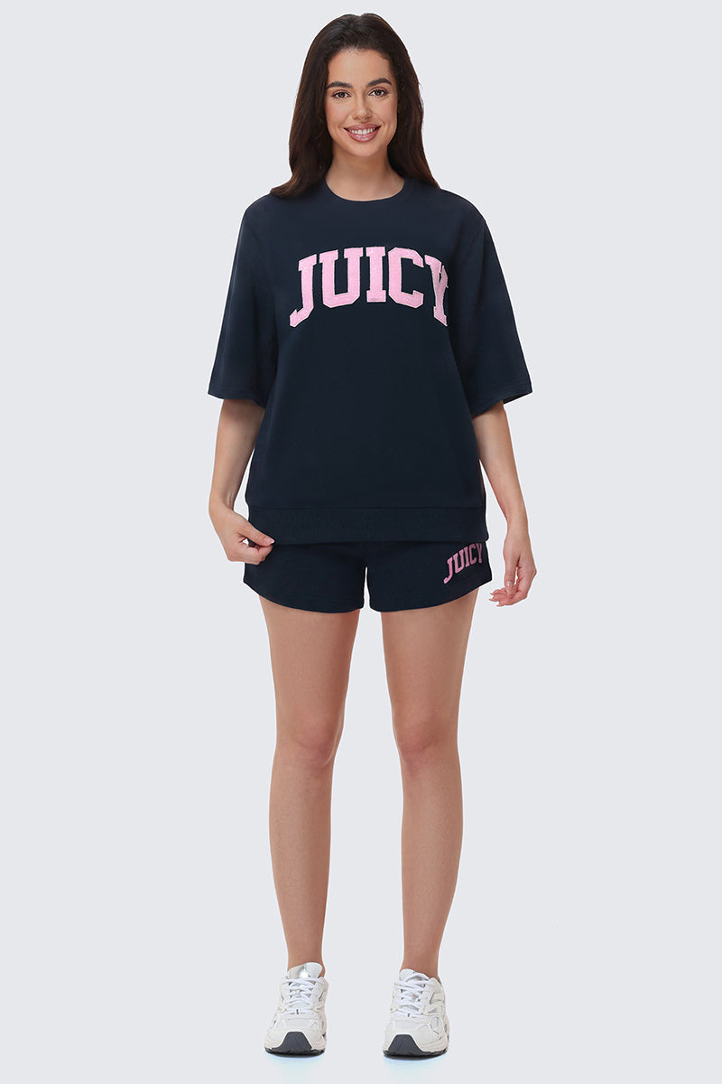 A woman smiles facing forward in a black Collegiate Sweatshirt and matching shorts, both with large pink JUICY lettering. She pairs the comfy set with white sneakers, posing against a plain light background.