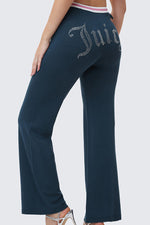 A woman wears Big Bling Wide Leg Pants in dark blue velour, featuring a rhinestone Juicy logo on the back and a pink-and-white contrast striped waistband, styled with high-heeled sandals.