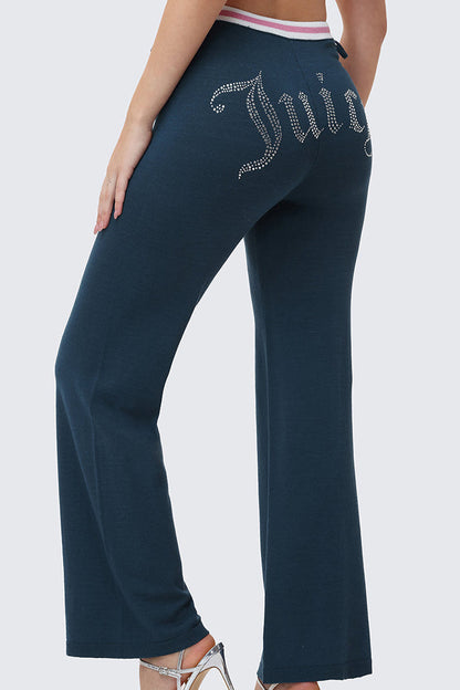 A woman wears Big Bling Wide Leg Pants in dark blue velour, featuring a rhinestone Juicy logo on the back and a pink-and-white contrast striped waistband, styled with high-heeled sandals.