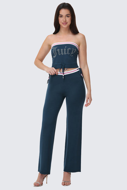A woman smiles at the camera in heels, wearing Big Bling Wide Leg Pants—a strapless, dark teal two-piece with the Juicy logo on top, contrast striped waistband, white and pink trim, drawstring waist, and ribbed wide-leg pants.