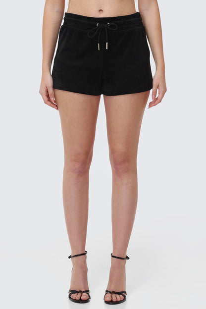 A woman stands against a light gray background, wearing Big Bling Velour Shorts with a drawstring waist and black strappy high-heeled sandals. The image is cropped from the waist to the feet.