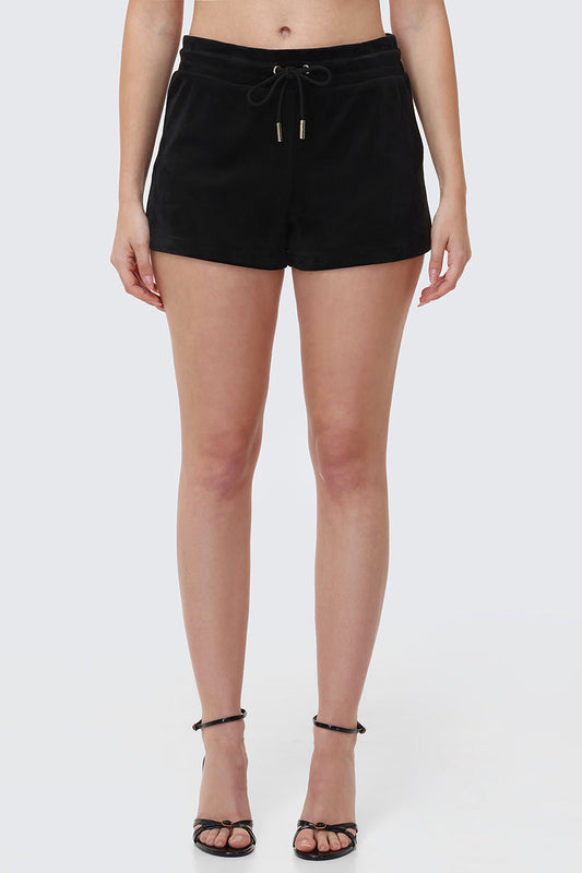 A woman stands against a light gray background, wearing Big Bling Velour Shorts with a drawstring waist and black strappy high-heeled sandals. The image is cropped from the waist to the feet.