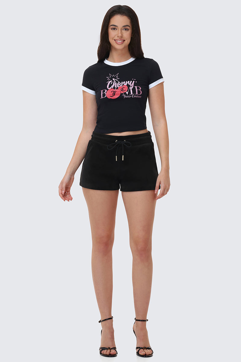 A woman smiles in a black crop top with Cherry Bomb and cherry graphics, paired with Big Bling Velour Shorts featuring a drawstring waist and black heeled sandals, set against a plain light background.