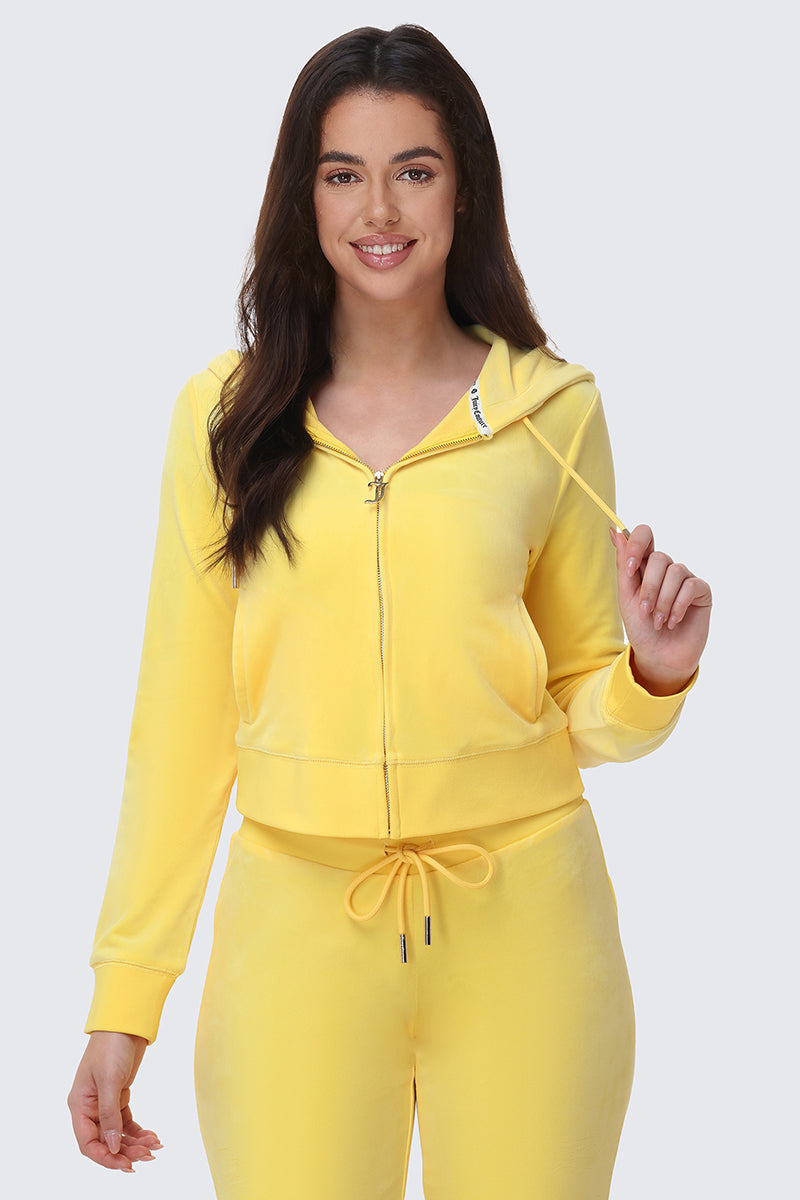 A woman with long dark hair smiles while wearing the OG Big Bling Velour Hoodie in bright yellow, paired with matching sweatpants. She holds the hoodie’s drawstring against a plain light gray background.