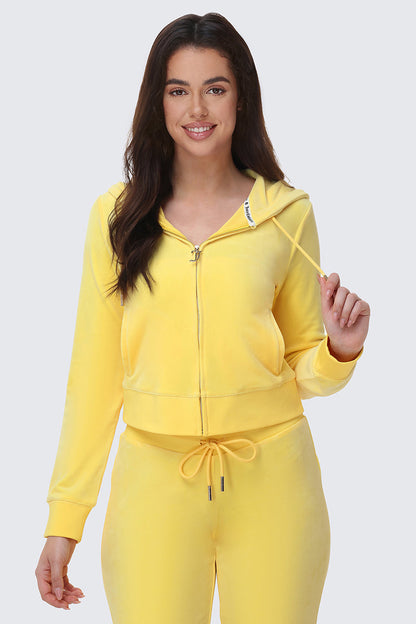 A woman with long dark hair smiles while wearing the OG Big Bling Velour Hoodie in bright yellow, paired with matching sweatpants. She holds the hoodie’s drawstring against a plain light gray background.