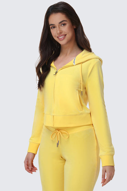 A woman with long dark hair smiles while wearing the OG Big Bling Velour Hoodie and matching drawstring sweatpants in yellow, standing against a plain light gray background.