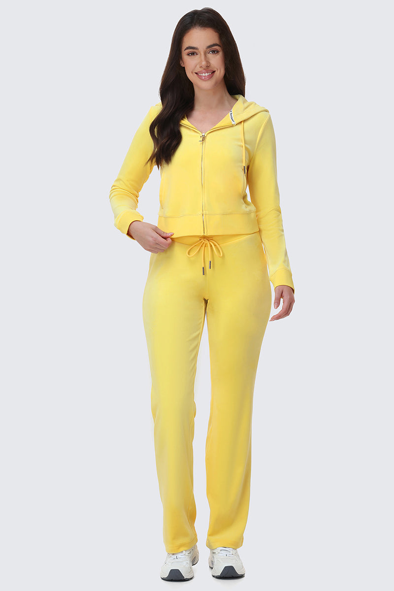 A woman smiles, wearing the OG Big Bling Velour Hoodie in bright yellow with matching drawstring pants and white sneakers, standing against a plain light gray background.