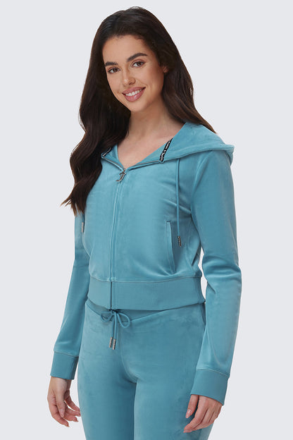 A woman with long brown hair wears the OG Big Bling Velour Hoodie in light blue, standing and smiling at the camera against a plain light background.