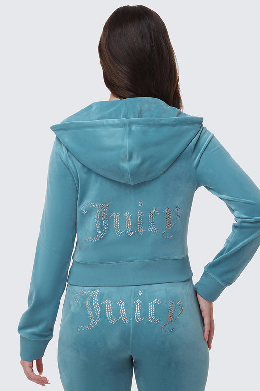 A person with long brown hair wears a light blue OG Big Bling Velour Hoodie and matching tracksuit, both featuring sparkling "Juicy" rhinestone embellishments on the back.