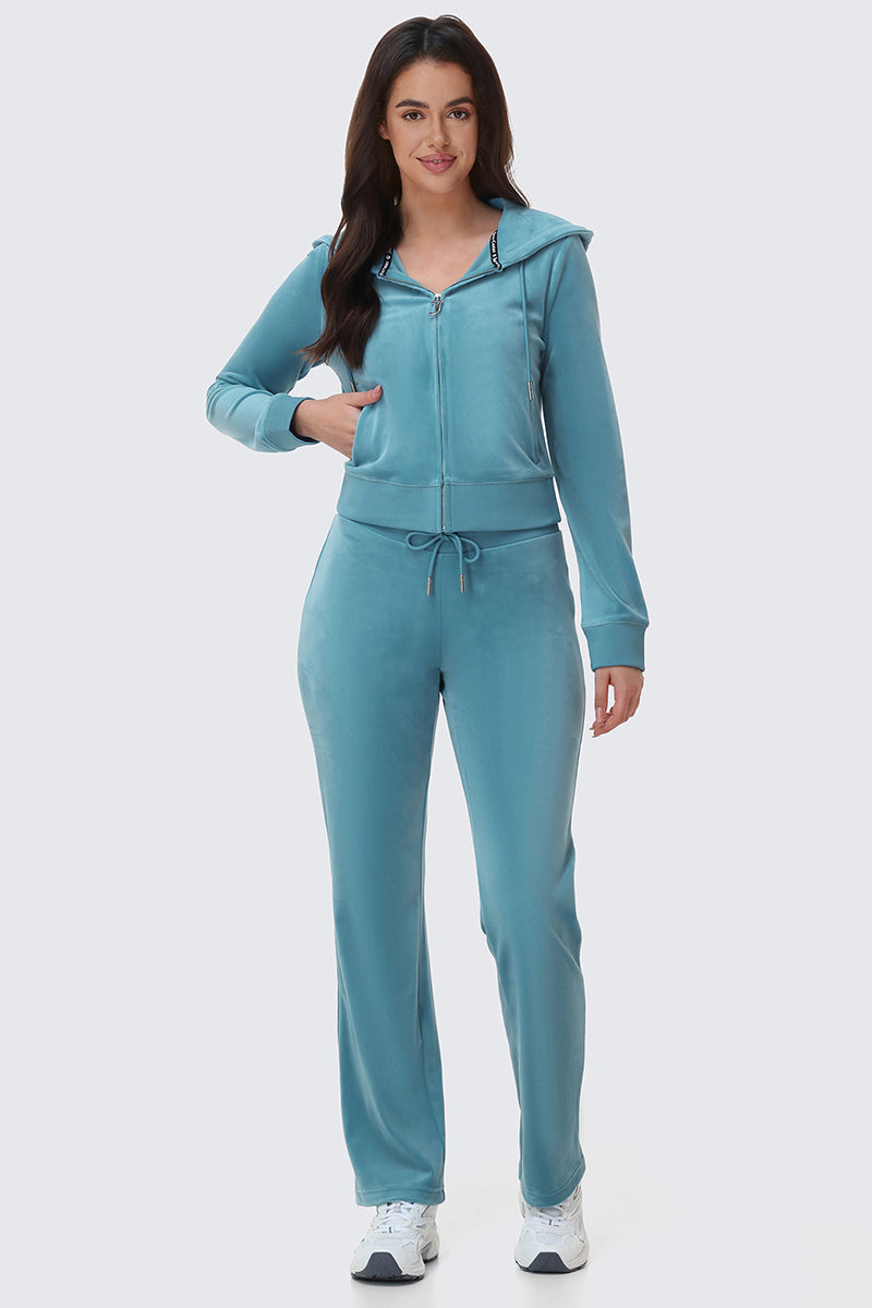 A woman with long dark hair smiles in the OG Big Bling Velour Hoodie and matching drawstring pants, paired with white sneakers. She poses against a plain light background.