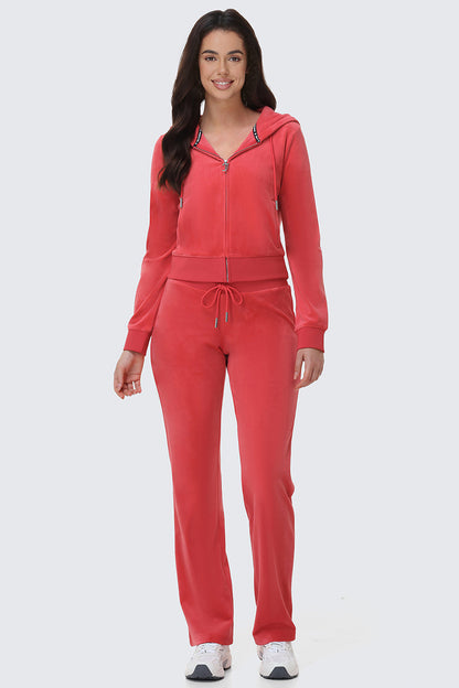 A woman with long dark hair smiles, wearing the OG Big Bling Velour Hoodie in bright pink, paired with matching straight-leg pants and white sneakers. The zip-up hoodie features a drawstring waist. She stands against a plain light gray background.