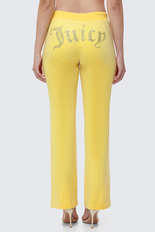 A woman shows off Y2K style in OG Big Bling Velour Track Pants with “Juicy” in rhinestones on the back, posing with her back to the camera and pairing the look with high-heeled sandals.