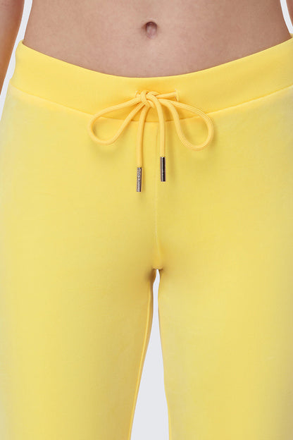 Close-up of a person wearing bright yellow OG Big Bling Velour Track Pants with a drawstring bow at the waistband, inspired by iconic Y2K style. The background is plain and light-colored.