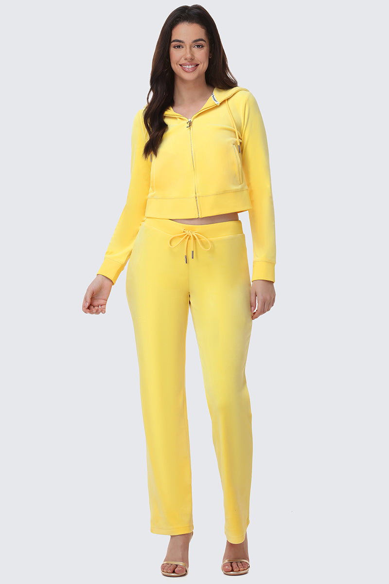 A smiling woman in OG Big Bling Velour Track Pants and a bright yellow Juicy zip-up hoodie stands facing forward, one hand by her side and the other slightly raised, finishing her Y2K look with open-toe heels.