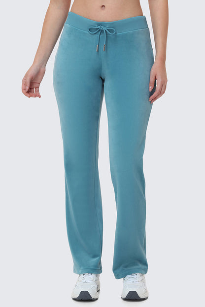 A person wearing the OG Big Bling Velour Track Pants in teal with white sneakers, standing against a plain light gray background. The image is cropped from shoulders to feet.