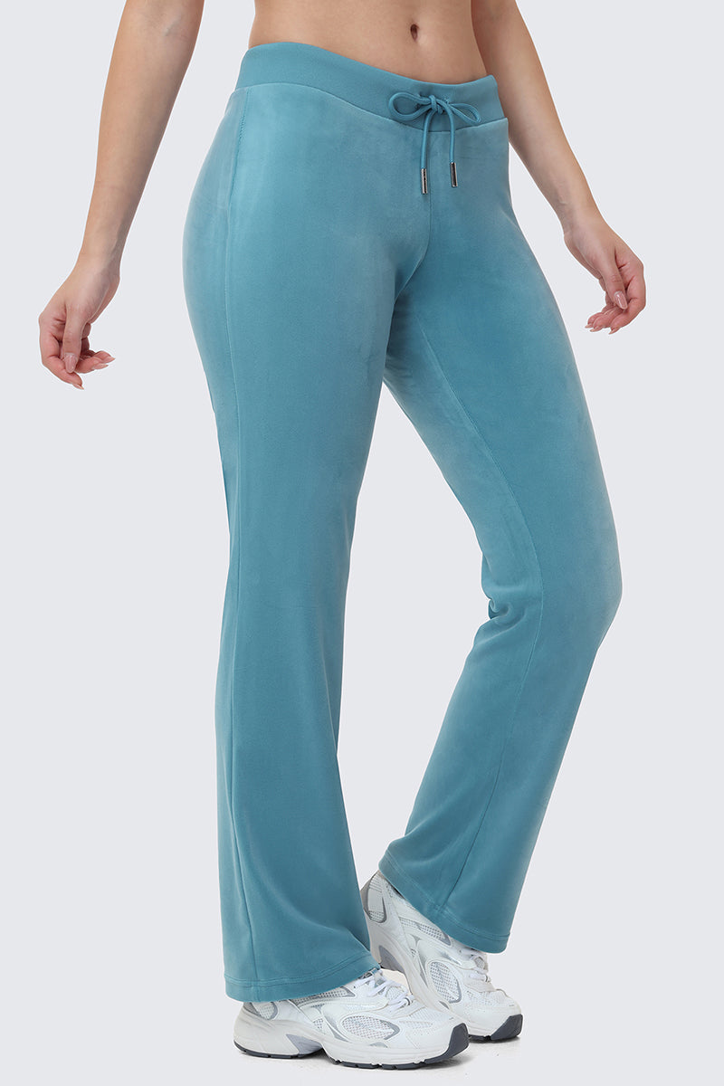 A person wears OG Big Bling Velour Track Pants in teal with a drawstring waist and white sneakers, standing against a light gray background. Only the lower half is shown, giving this Y2K-inspired look a luxe twist.