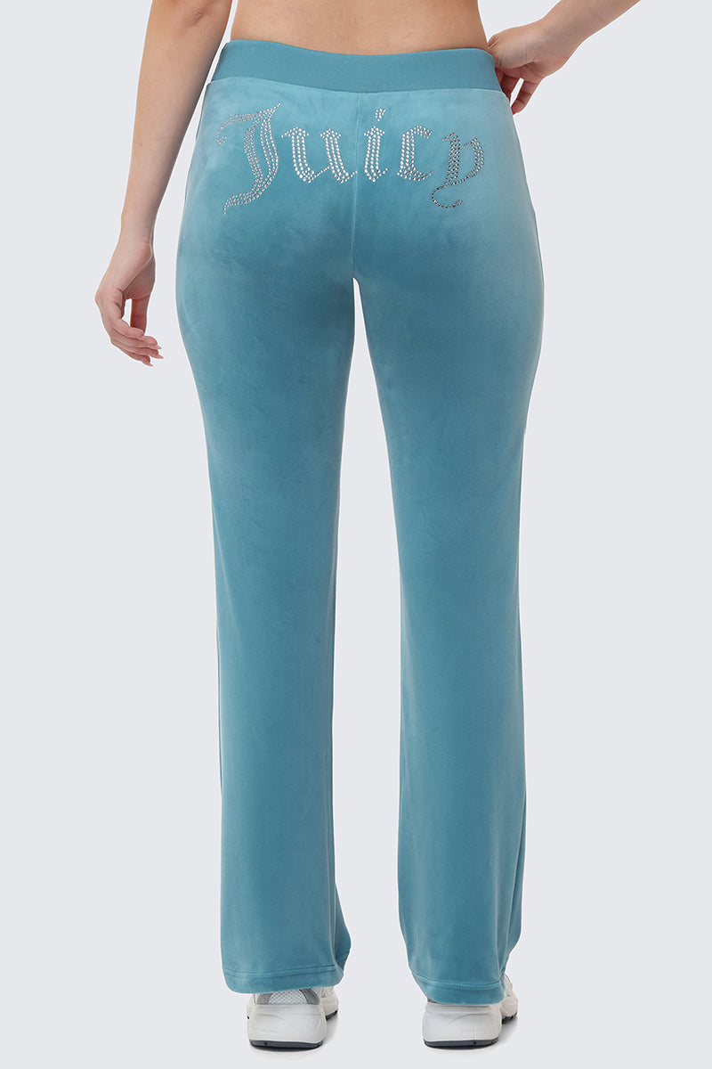 A person stands with one hand on their hip, shown from behind in light blue OG Big Bling Velour Track Pants featuring a Juicy rhinestone embellishment across the back, paired with white shoes.