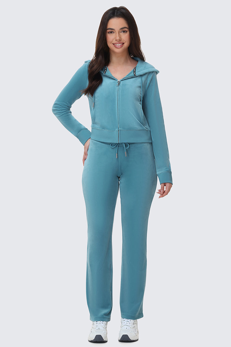 A woman with long dark hair smiles while wearing OG Big Bling Velour Track Pants, a matching teal zip-up hoodie, and white sneakers. She stands against a plain light gray background.