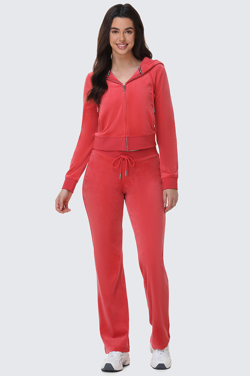 A woman smiles while wearing bright red OG Big Bling Velour Track Pants with a zip-up hoodie and white sneakers, channeling iconic Y2K style against a plain light gray background.