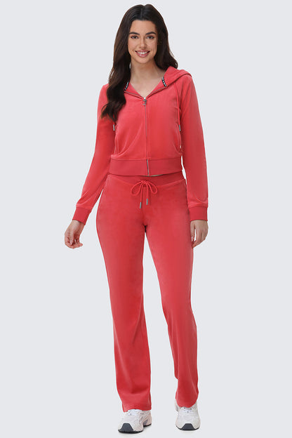 A woman smiles while wearing bright red OG Big Bling Velour Track Pants with a zip-up hoodie and white sneakers, channeling iconic Y2K style against a plain light gray background.