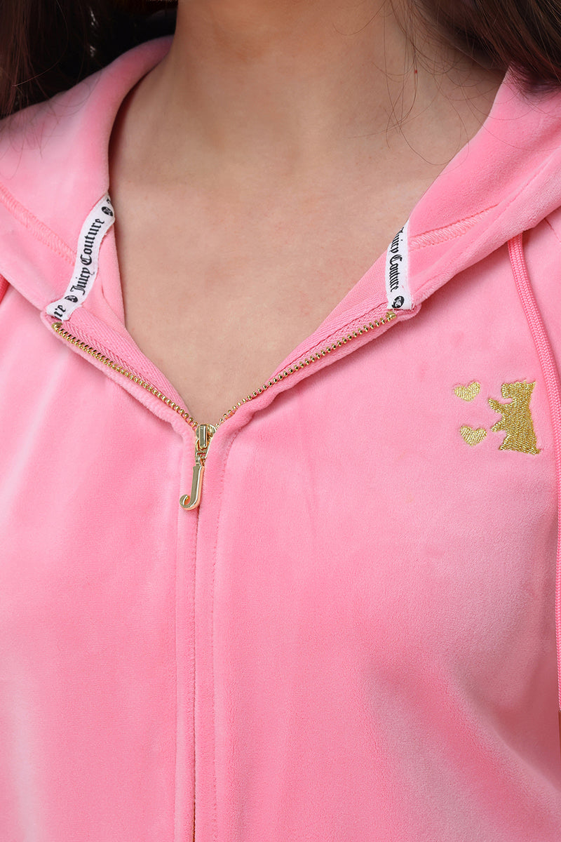 Close-up of a person wearing the Juicy Couture Heritage Stripe Bling Velour Zip Hoodie in light pink, featuring a gold crown logo on the left side and branded text on the inner hood. The soft velour fabric looks plush and luxurious.