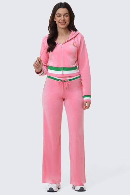 A woman smiles in the Juicy Couture Heritage Stripe Bling Velour Zip Hoodie and matching drawstring pants with green and white striped bands, rhinestone logo, and white sneakers, against a plain light gray background.
