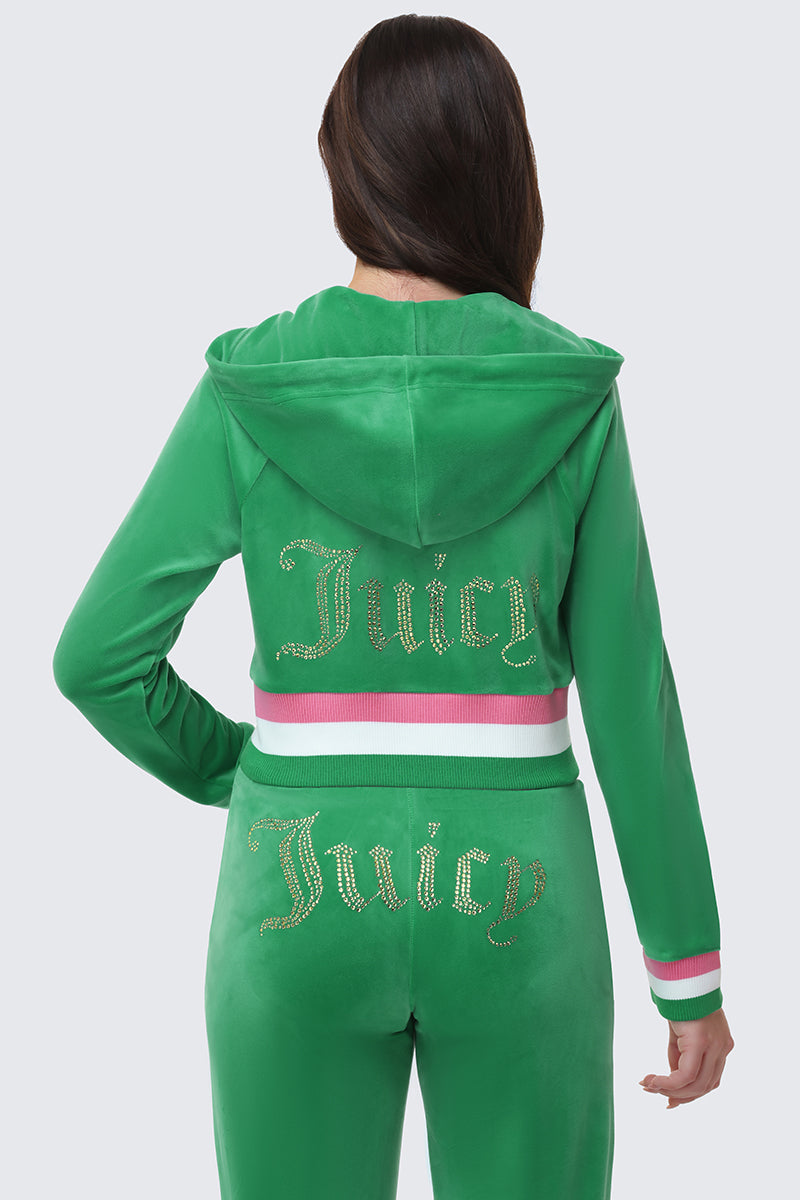 A woman is seen from behind wearing the green Heritage Stripe Bling Velour Zip Hoodie, featuring pink and white striped trim and a gold rhinestone logo on the back, paired with matching velour pants.