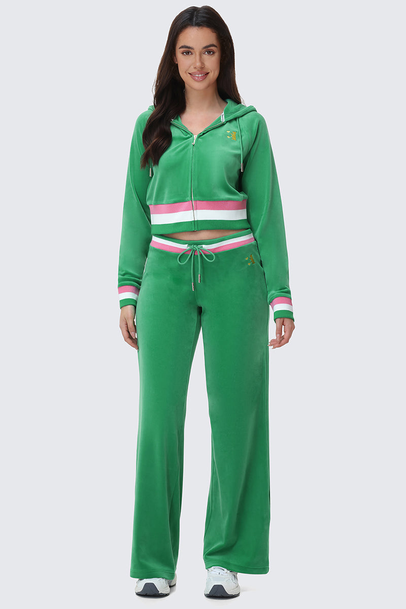 A woman faces forward wearing the Heritage Stripe Bling Velour Zip Hoodie by Juicy Couture, featuring a green velour finish, white/red/green striped waistband and cuffs, rhinestone logo, drawstring hood, and paired with white sneakers.