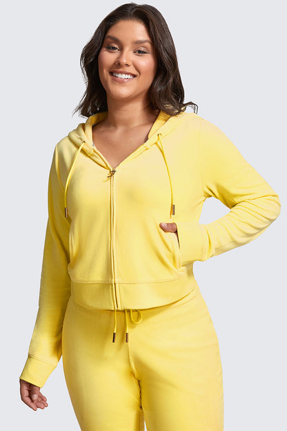 A woman smiles while wearing the Plus-Size OG Big Bling Velour Hoodie in bright yellow with matching pants, one hand in her pocket, bringing a luxe vibe to her look against a light gray background.
