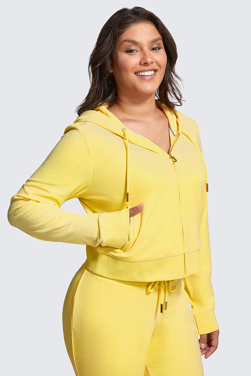 A woman smiles wearing the Plus-Size OG Big Bling Velour Hoodie in yellow with matching sweatpants, one hand in her pocket, standing against a light gray background for a luxe take on casual style.