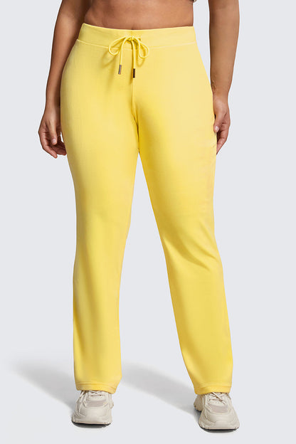 A person wearing Plus-Size OG Big Bling Velour Track Pants in bright yellow with white sneakers stands against a plain light grey background.