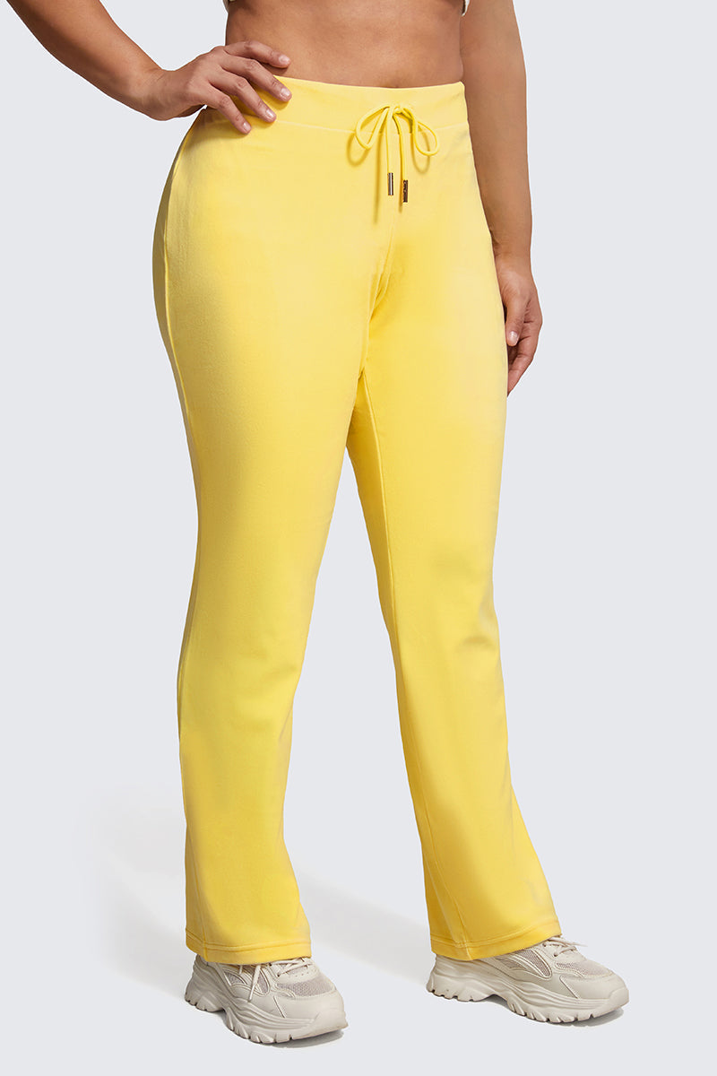 A person models the Plus-Size OG Big Bling Velour Track Pants in bright yellow, paired with white sneakers and posed with one hand on their hip against a light gray background—a stylish Y2K-inspired look.