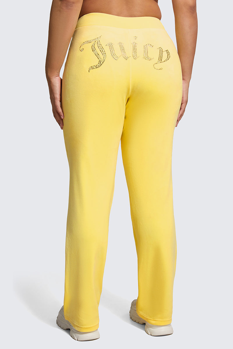A person is seen from behind wearing Plus-Size OG Big Bling Velour Track Pants in bright yellow with a sparkling Juicy embellishment across the back, styled with classic white sneakers for a Y2K-inspired look.