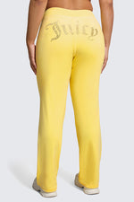 A person is seen from behind wearing Plus-Size OG Big Bling Velour Track Pants in bright yellow with a sparkling Juicy embellishment across the back, styled with classic white sneakers for a Y2K-inspired look.