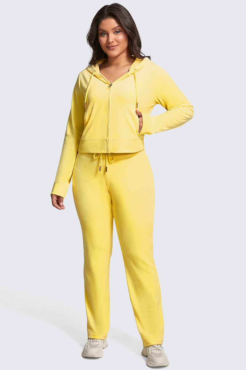 A woman smiles in Plus-Size OG Big Bling Velour Track Pants, styled with a matching yellow velour zip-up hoodie and white sneakers. Her long, dark hair and the light gray background highlight her chic Y2K-inspired look.