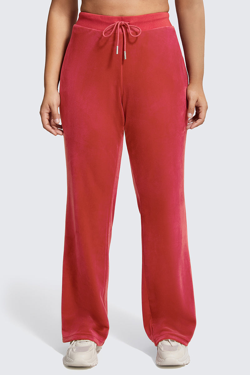 Wearing the Plus-Size OG Big Bling Velour Track Pants in red with a drawstring waist and subtle Juicy embellishment, paired with white sneakers, a person stands against a plain light background.