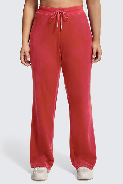 Wearing the Plus-Size OG Big Bling Velour Track Pants in red with a drawstring waist and subtle Juicy embellishment, paired with white sneakers, a person stands against a plain light background.