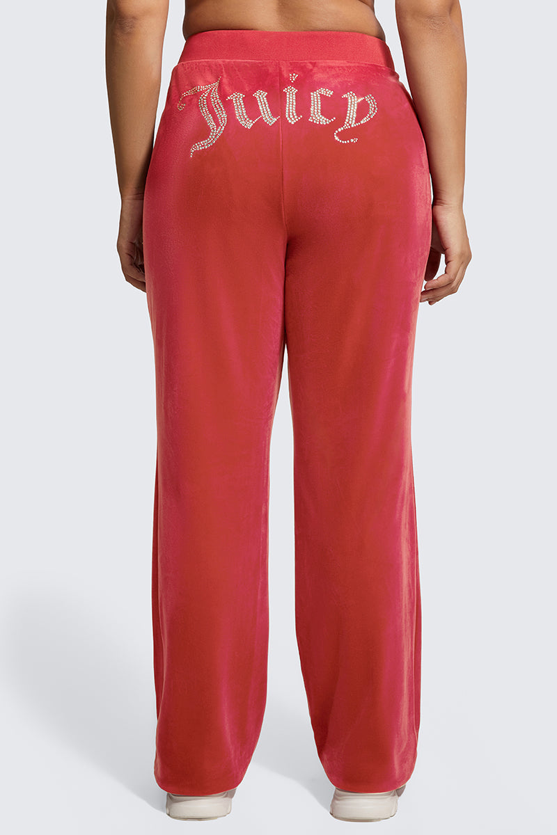A person stands with their back to the camera against a plain light background, wearing Plus-Size OG Big Bling Velour Track Pants featuring Y2K-inspired red velour and a sparkling Juicy rhinestone embellishment on the back.