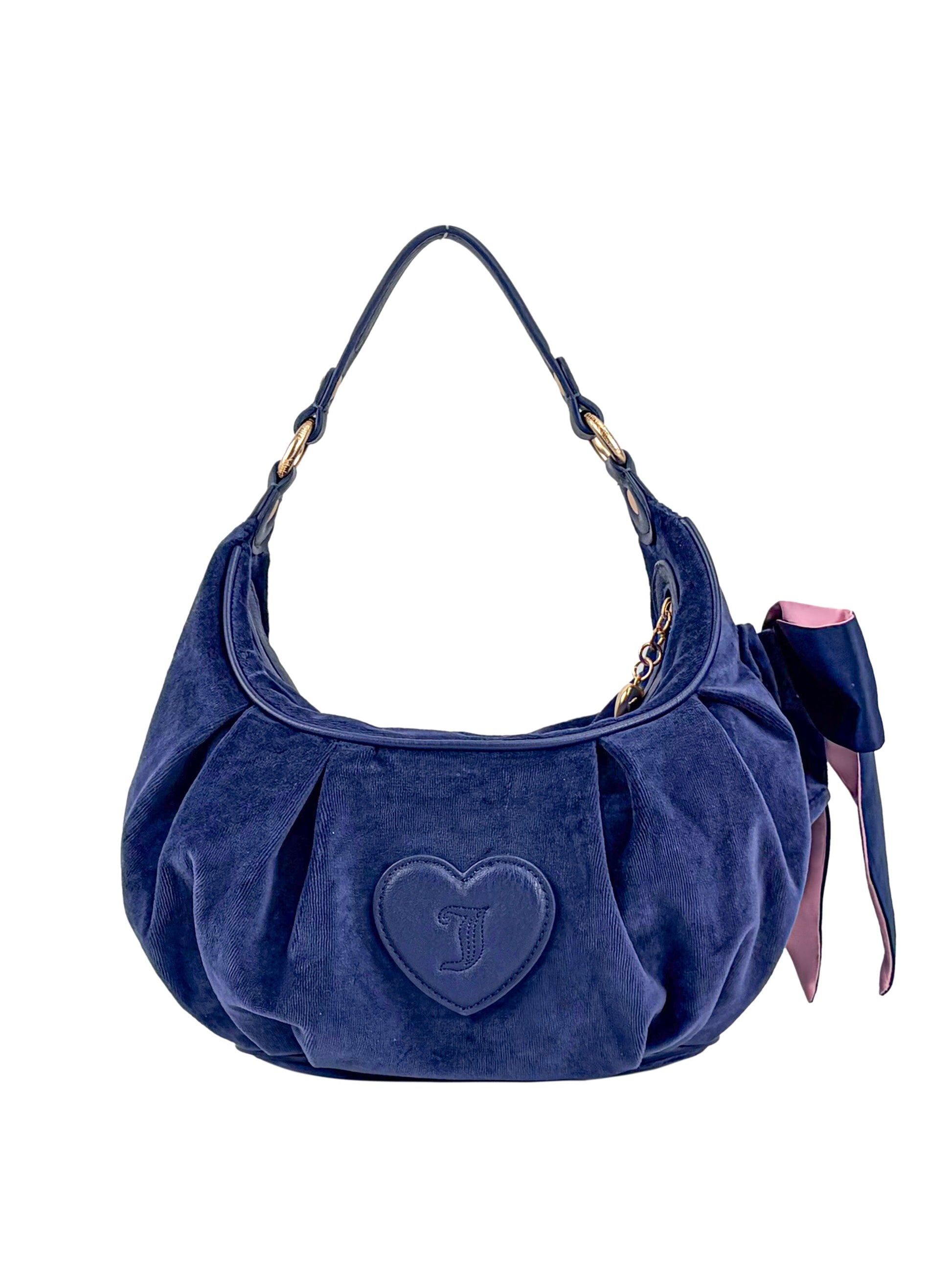 The Royal Bloom Hobo Bag is a dark blue velvet handbag featuring a curved handle, gold hardware, and a large heart-shaped stitch on the front. A pink and navy ribbon tied to the handle adds a feminine accent.