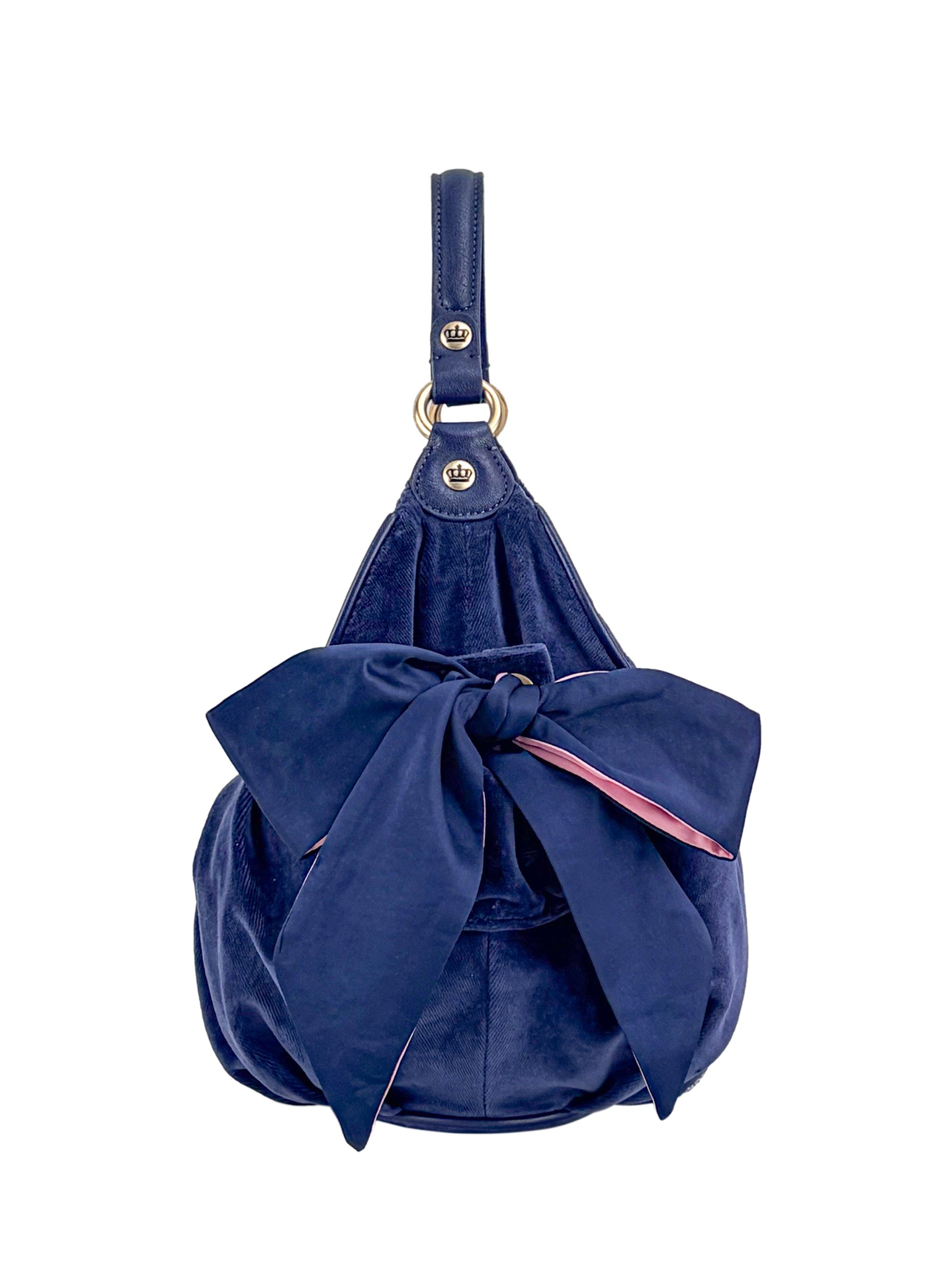 The Royal Bloom Hobo Bag is a navy blue handbag with a large matching front bow and a short handle with metal rings, showcased against a white background.