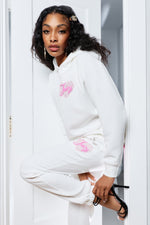 A woman with long dark hair and hair clips poses in a doorway wearing the Sweetheart Fleece Oversized Hoodie and matching sweatpants with pink designs, paired with black high-heeled sandals for a chic yet casual look.