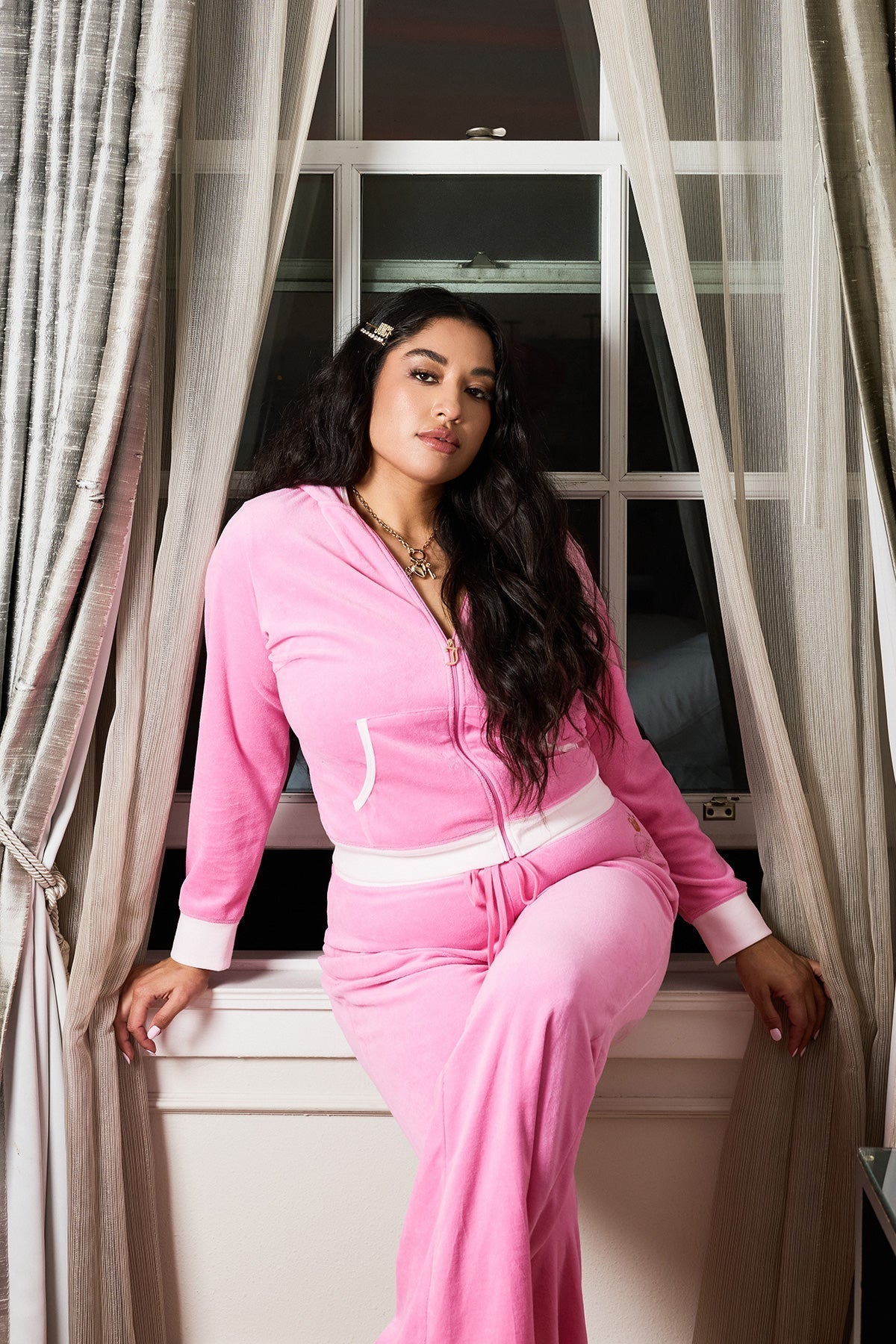 A woman with long dark hair wears a Plus-Size Lady Juicy Hollywood Heritage Hoodie in pink velour as she sits on a windowsill between open beige curtains, confidently gazing at the camera.
