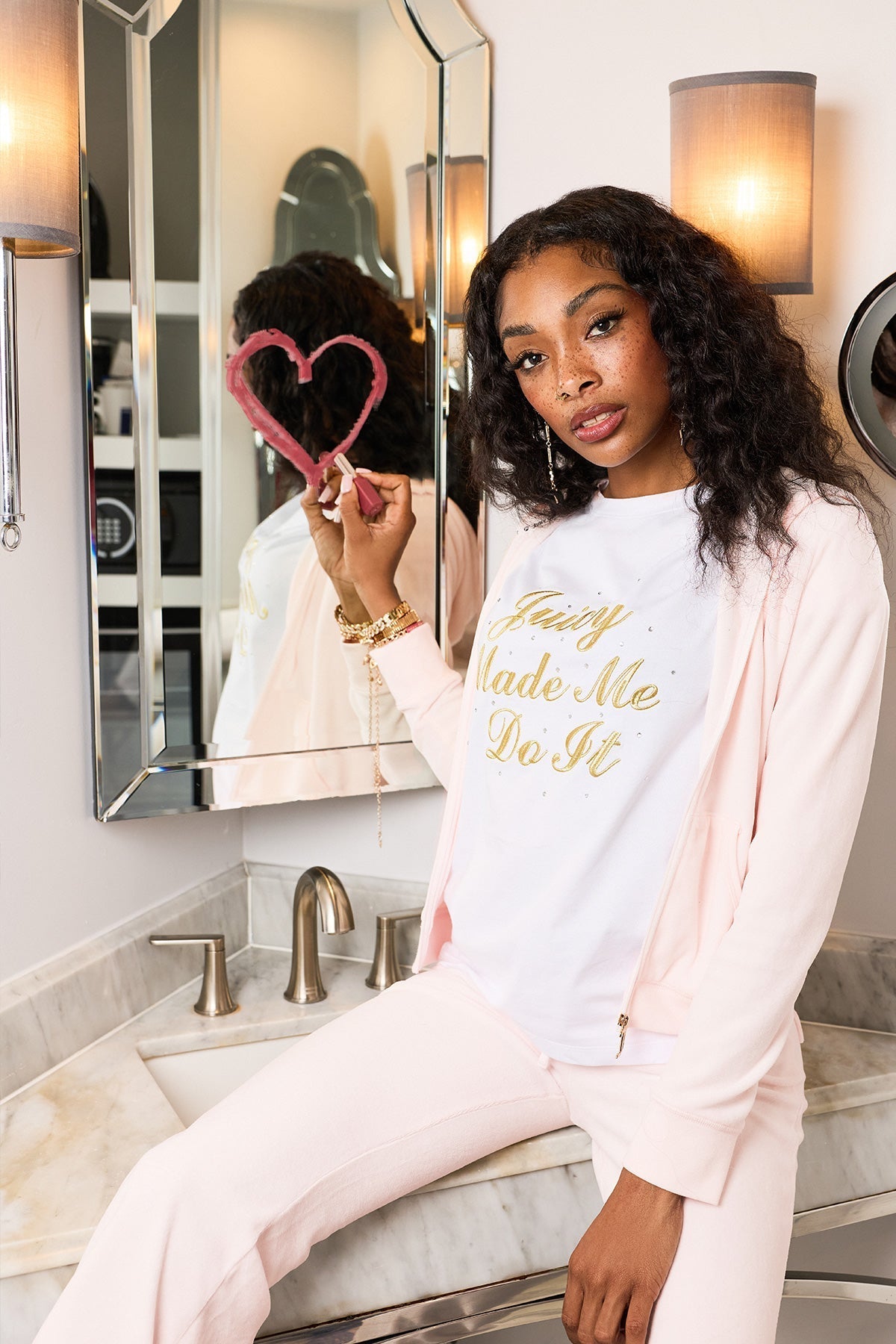 A woman in a white and light pink outfit sits on a bathroom counter, holding up a heart drawn on the mirror with lipstick. She wears the Made Me Do It Heritage Oversized Tee, making it the statement piece as she softly gazes at the camera.