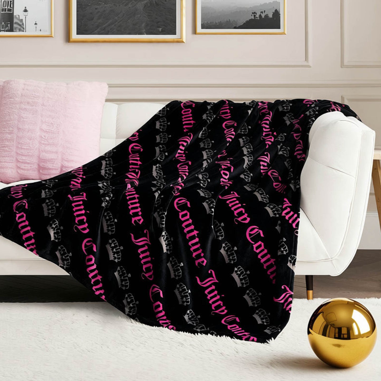 A white sofa with a pink pillow and the Plush Throw Blanket, featuring pink text and crowns, draped over it. A gold decorative ball rests on a beige rug in front, while framed pictures are displayed on the wall behind.