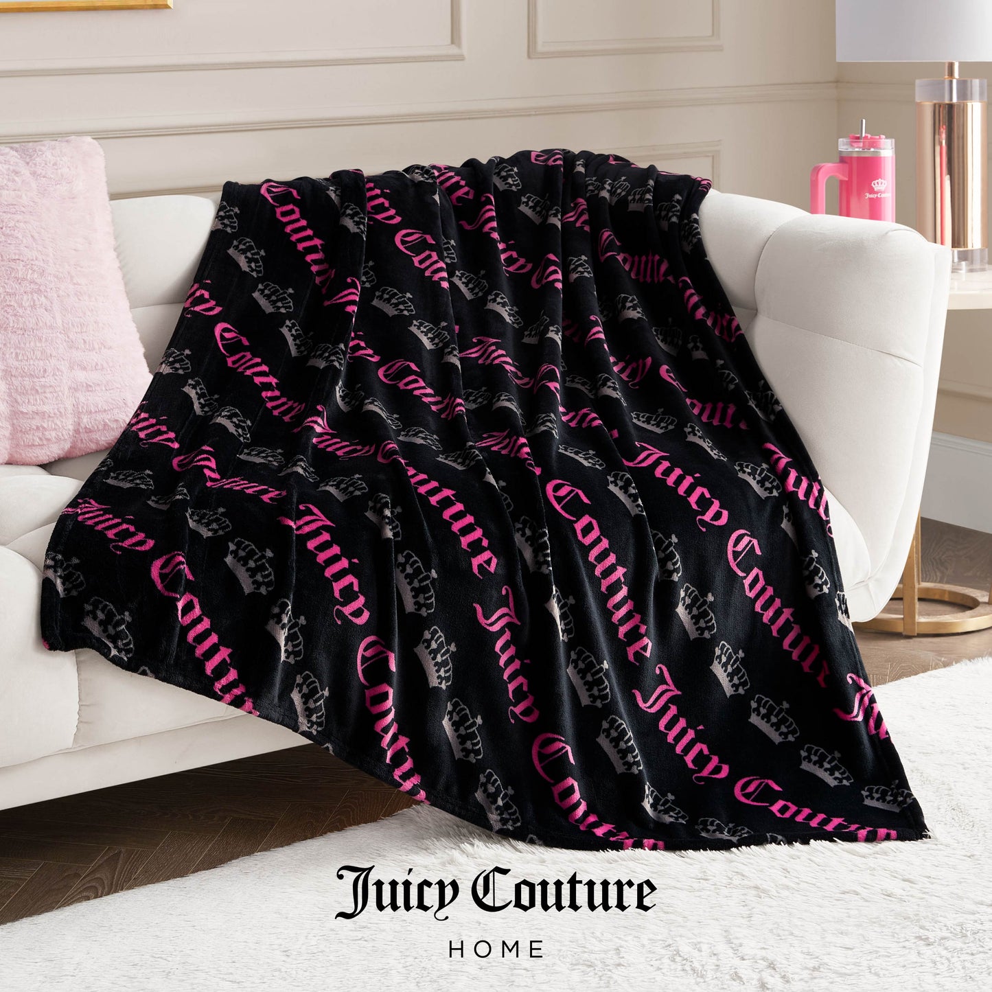 A Plush Throw Blanket with black fabric, pink Juicy Couture lettering, and crown logos is draped over a white sofa in a cozy modern living room with pink accents, adding an ultra-soft, stylish touch to the inviting space.