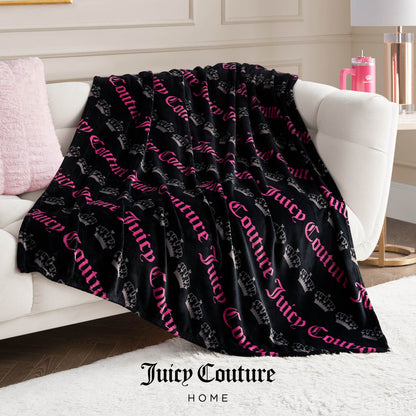 A Plush Throw Blanket with black fabric, pink Juicy Couture lettering, and crown logos is draped over a white sofa in a cozy modern living room with pink accents, adding an ultra-soft, stylish touch to the inviting space.