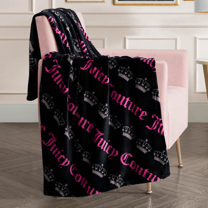 The Plush Throw Blanket, ultra-soft and plush, showcases gray crown patterns with bold pink text. Draped over a light pink armchair, it enhances the modern room’s style with wood floors and white paneled walls.