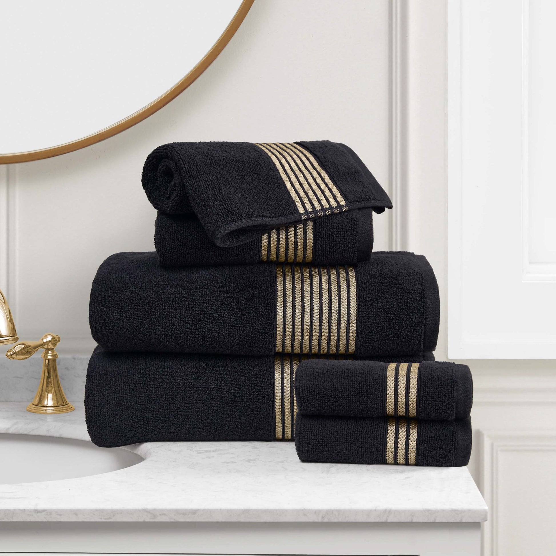 A stack of Melissa Bath Towel Set, made from 100% cotton towels with gold stripes, is neatly arranged on a white marble bathroom countertop beside a gold faucet and mirror.